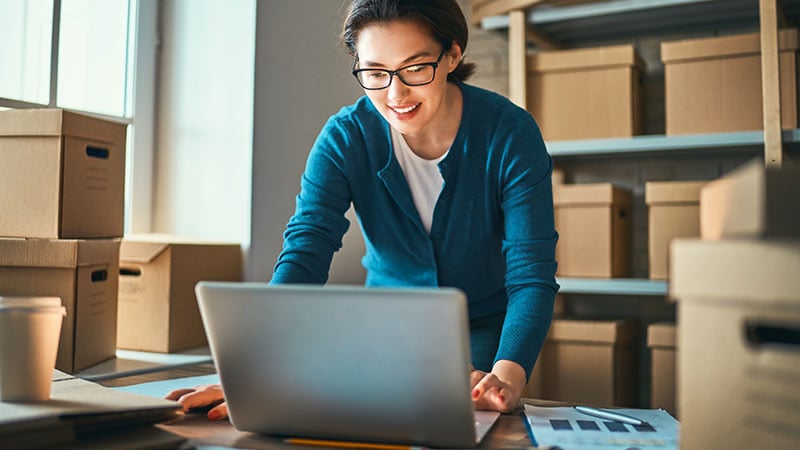 Mulher de óculos, em pé, cercada de caixas de papelão, sorrindo para o notebook